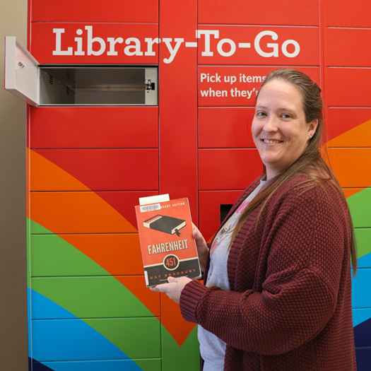 Smiling person holding a book in front of a Library-To-Go locker