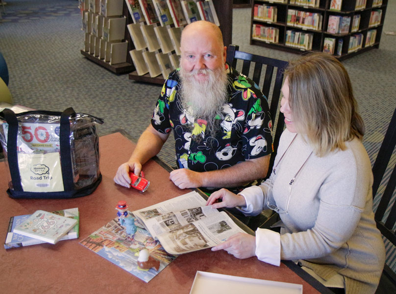 And older person and a younger one sit together at a table examining items from a library Memory Care Kit