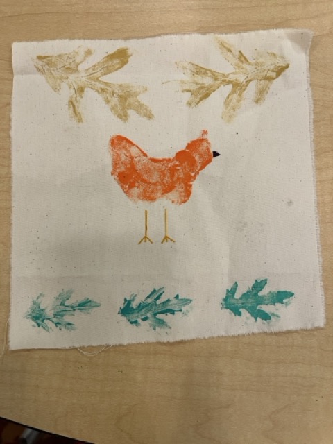 A sample nature print on a scrap of off-white speckled fabric. In the center of the fabric scrape is an orange chicken with yellow legs and dark purple beak. At the top of the fabric in each corner are oak leaves printed in gold paint. Running across the bottom of the fabric scrap are three mum leaves printed in green. 