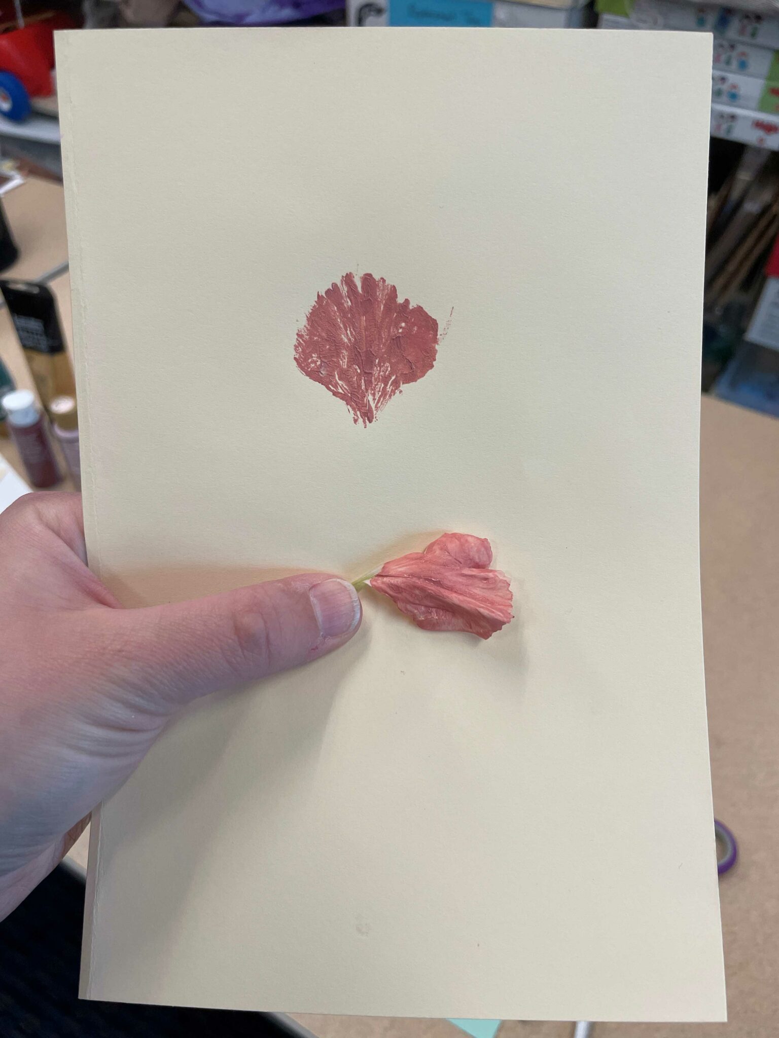 A rectangular piece of cream-colored cardstock with a carnation petal printed on it around the top middle. The painted petal print is a shade of rose pink. Below the printed petal is a white thumb holding the live petal that was used to make the print. 