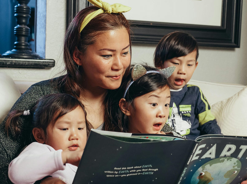 A family consisting of a mom and three young kids sits on a couch looking at an open picture book called "Earth." One child looks very excited.