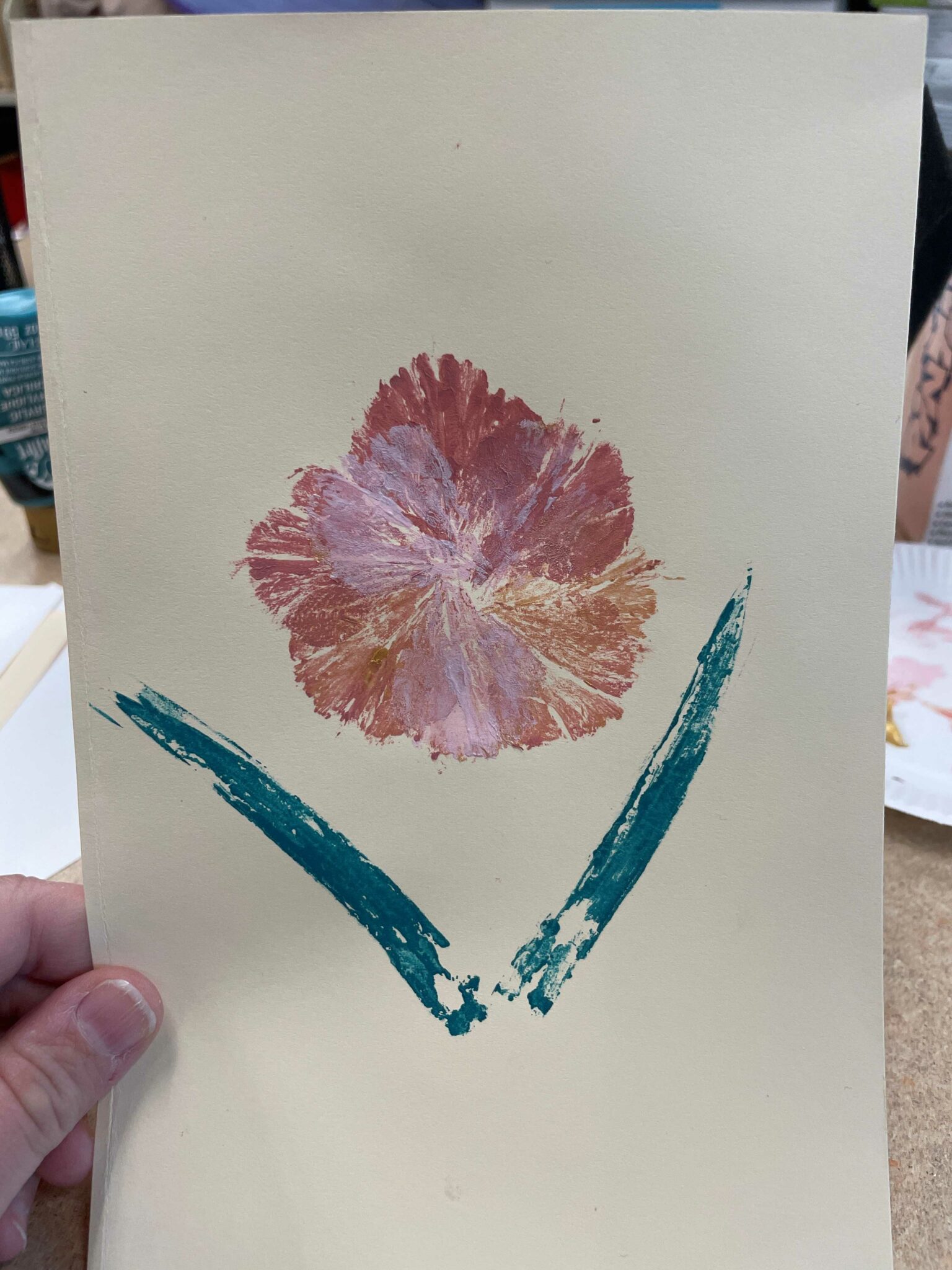 A rectangular piece of cream-colored cardstock with a carnation flower printed in the middle. The printed flower is made up of a single carnation petal painted and then pressed to the paper eight times in a circular overlapping pattern. Beneath the flower head (made up of printed petals) are two carnation leaves printed in a dark blue green paint. The leaves are arranged below the flower head in a loose V shape. 