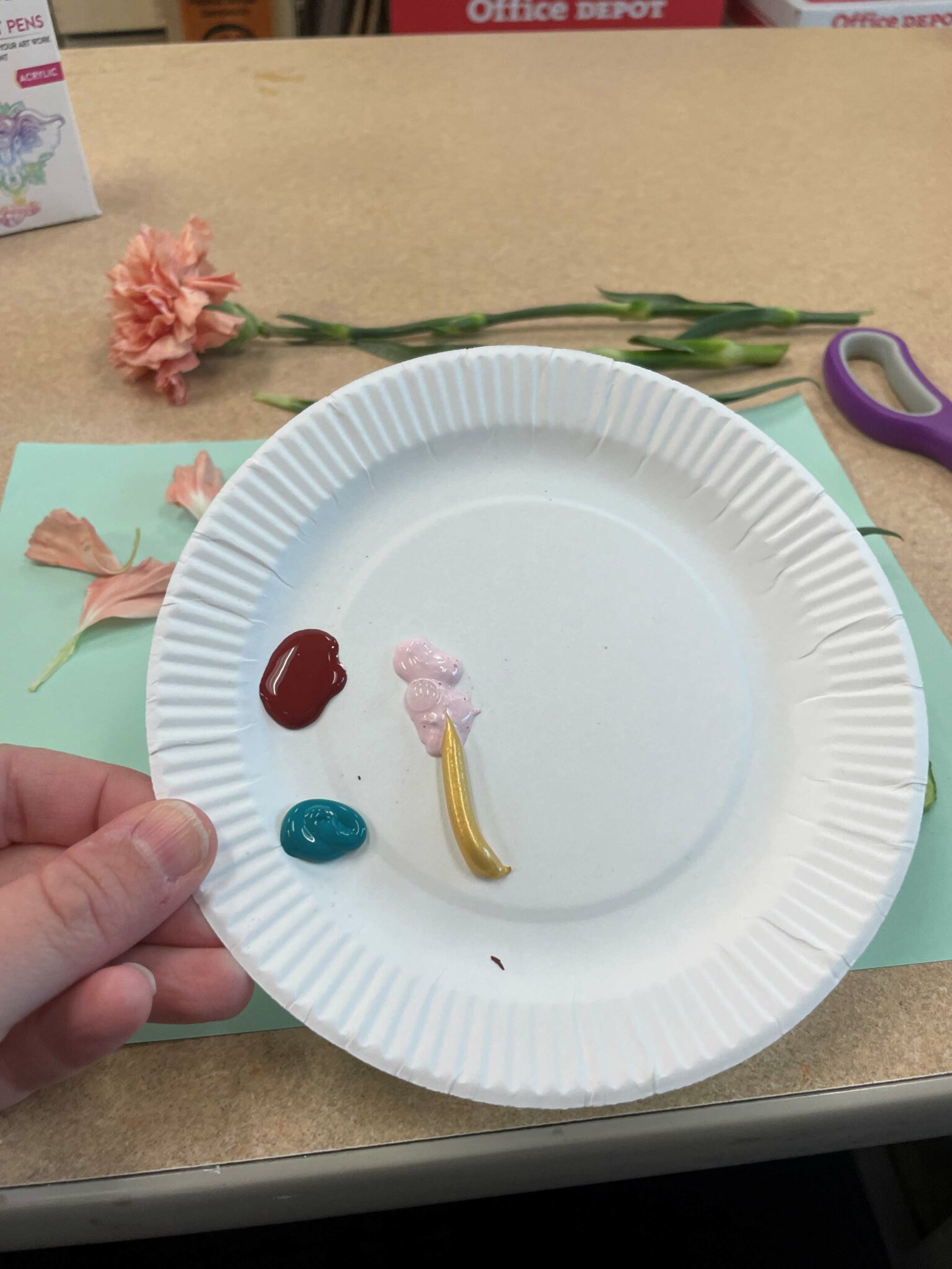A close-up of a paper plate palette with four small blobs of paint on it in a dark red shade, a light pink shade, a gold shade, and a dark blue green shade. 