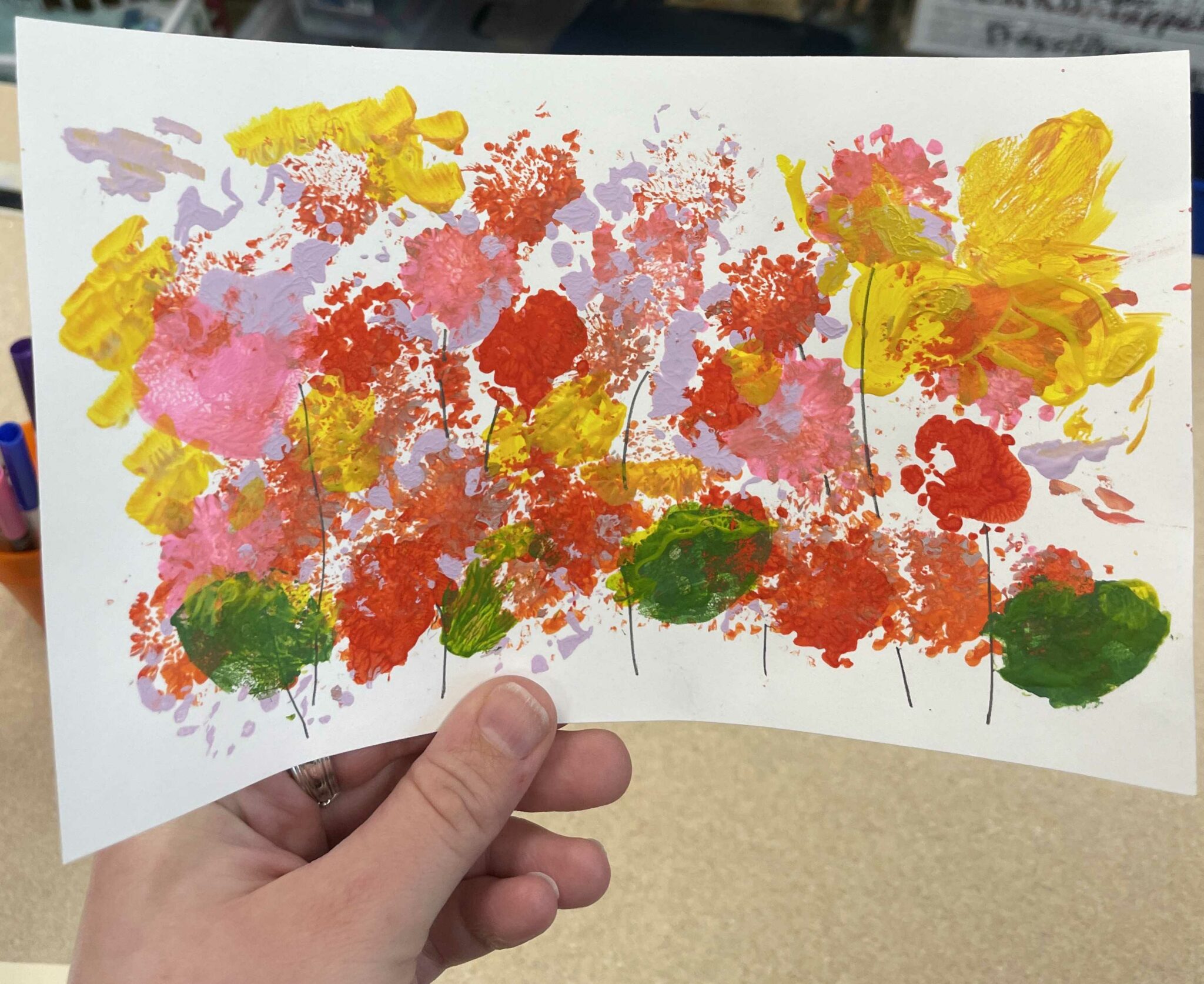 A sample nature print featuring lilac, yellow, pink, red, orange and green blotches of paint smattered all over a piece of white rectangular cardstock. Amidst the colorful jumble of abstract paint marks are 10ish lines drawn in pencil to suggest stems to the brightly colored paint jumbles. 