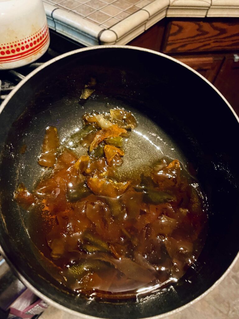 Photo of a black saucepan filled with candied citrus peels and syrup. The peels' colors (green, yellow, orange and reddish orange) have become muted in the dark amber-colored syrup.