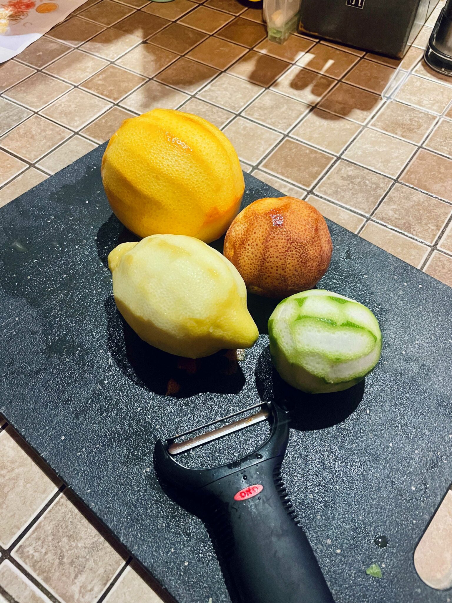 Clockwise from top left: a Cara Cara orange, a blood orange, a lime and a lemon rest on a silver-speckled black cutting board. The peel of each fruit has been removed in strips with a black vegetable peeler, which rests on the cutting board below the fruits in the bottom center of the photo.