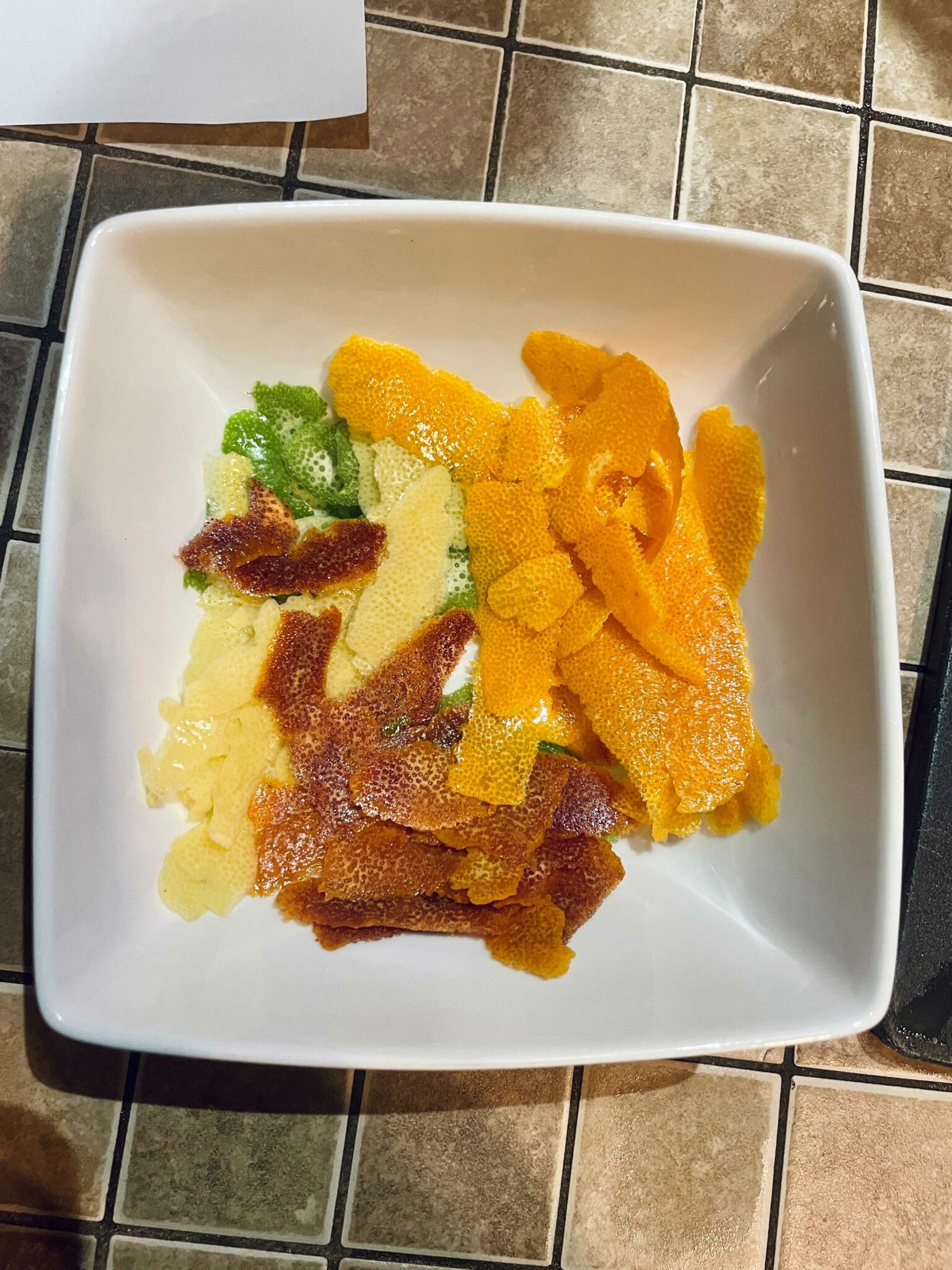 A square white bowl with strips of citrus peel inside it. The peels are orange, reddish orange, yellow and green.