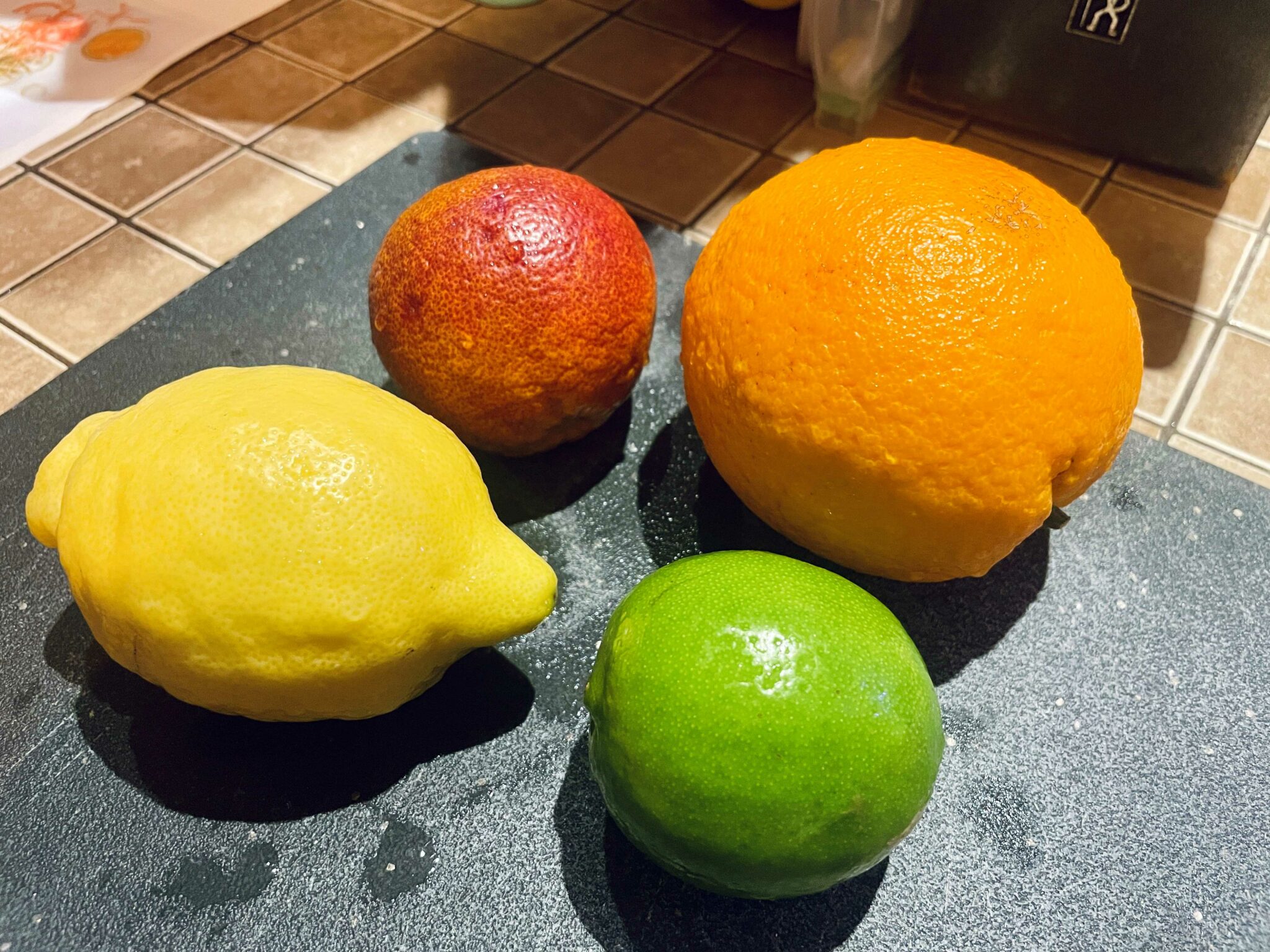 Clockwise from top left: a blood orange, a Cara Cara orange, a lime and a lemon rest on a silver-speckled black cutting board.