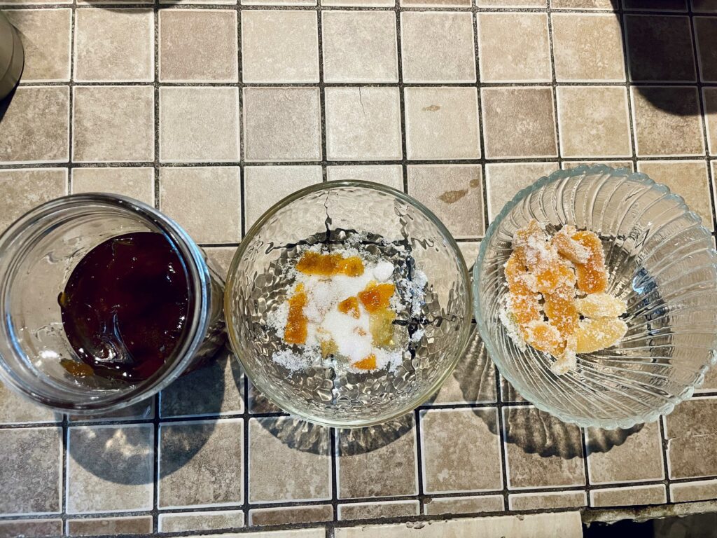 Photo of one 16 oz glass mason jar and two small glass bowls in a line on a taupe tiled counter. The glass jar contains 1/3 cup of candied citrus peel and syrup, which is a dark amber color. The first glass bowl contains some strips of candied peel scattered in a big clump of white sugar. The third bowl contains some strips of candied peel that have been rolled in sugar; the sugar-dusted candied peel rest in the bowl on their own.