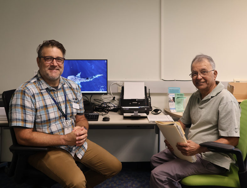 an instructor and a library patron with a folder of documents sit with a computer and scanner on a desk between them