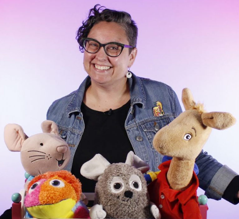 Hilary Aid, smiling and with arms out, stands behind several large puppets