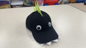 A children's craft project made from a black baseball cap sitting on a light-colored table. The cap is modified to resemble a dinosaur: two large plastic googly eyes are glued to the front panels, a row of white felt zig-zag teeth points outward from under the bill, and three stiff, lime-green foam spikes stick straight up along the center seam of the crown.