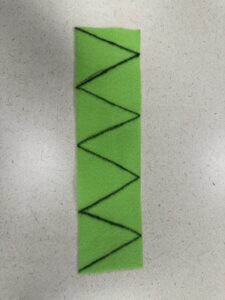 A rectangular strip of bright green felt lying vertically on a light surface, featuring a black zig-zag marker line drawn from top to bottom to create a template for cutting out triangular shapes.