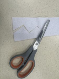 A pair of crafting scissors laying on a strip of white felt that has been cut into a zigzag, spiky pattern for teeth.