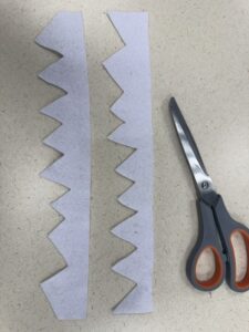 Two long, vertical strips of white felt cut into jagged, pointed dinosaur teeth patterns, laid next to a pair of crafting scissors.