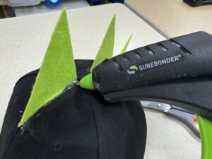 A hot glue gun applying adhesive to the base of a lime green felt triangle spike attached to a black cap.