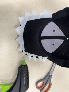 An overhead shot of a DIY dinosaur crafting project in progress. A black baseball cap sits upside down on a light-colored surface. A row of jagged, white triangle teeth cut out of felt has been glued along the inside curve of the hat's visor.