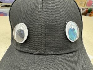 A close-up front view of a black baseball cap with two large googly eyes attached to the front panels directly above the brim.