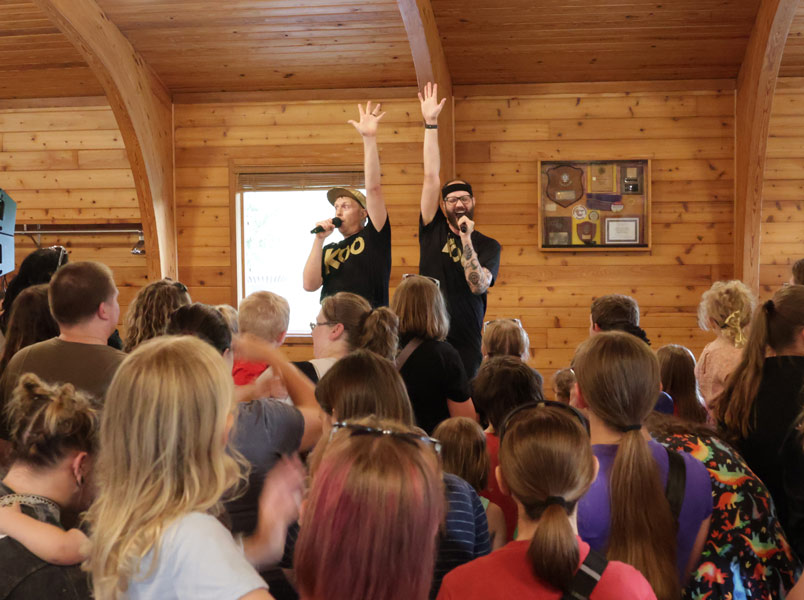The two members of Koo Koo sing into microphones and raise their hands in the air with a crowd of families watching in the foreground