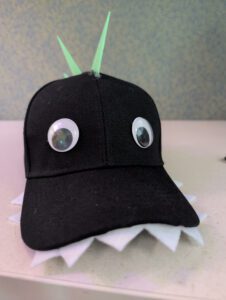 A children's craft project made from a black baseball cap sitting on a light-colored table. The cap is modified to resemble a dinosaur: two large plastic googly eyes are glued to the front panels, a row of white felt zig-zag teeth points outward from under the bill, and three stiff, lime-green foam spikes stick straight up along the center seam of the crown.