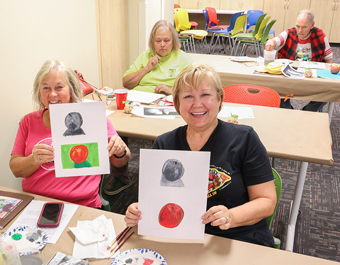 senior students in "The Creative Age" program seated at tables with art supplies hold up some of their painting exercises