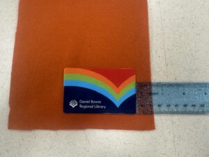 Sizing orange felt fabric using a library card and a clear ruler to measure seam allowance for a DIY card sleeve.