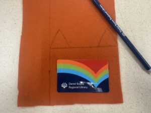 Tracing triangular fox ears onto orange craft felt with a pencil, using a library card to gauge the holder's width and height.