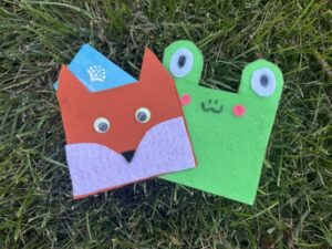 Two handmade felt animal card holders shaped like a fox and a frog, resting on green grass with a blue card tucked inside the fox holder.